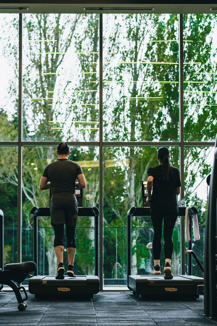 Two fitness enthusiasts running on treadmills in a gym with outdoor view.